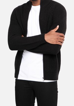 Zwarte gebreide cardigan met een gestructureerd, geribbeld ontwerp, hoge kraag en ritssluiting; gedragen over een wit t-shirt en zwarte broek.