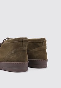 Botas de tobillo de ante verde oliva con suela de goma marrón, acentos cosidos y diseño minimalista. Presentan una puntera redonda y cordones en el costado.