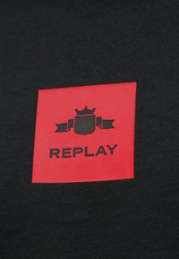 Κόκκινο τετράγωνο σε μαύρο ύφασμα με μαύρο έμβλημα κορώνας και ασπίδας πάνω από τη λέξη "REPLAY" με έντονα κεφαλαία γράμματα.