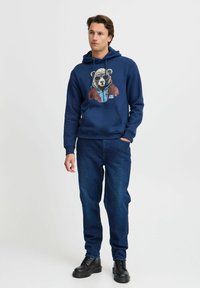 Felpa blu navy con grafica di un orso in occhiali da sole e giacca, abbinata a jeans di denim scuro e stivali neri.