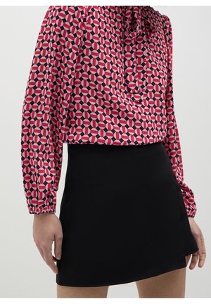 Femme portant un chemisier rose à manches longues avec un motif géométrique, rentré dans une mini-jupe noire unie, les mains détendues le long du corps.