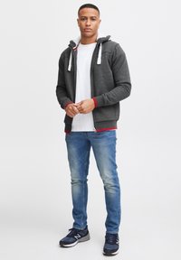 Grijze zip-up hoodie met witte voering, rode accenten bij de manchetten en zoom, gecombineerd met een wit t-shirt en blauwe spijkerbroeken. Zwarte sportschoenen.