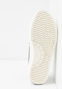 Skosula i vit gummi med horisontella spår och cirkulär logodetalj. Svart textilovandel synlig vid kanten av sulan.