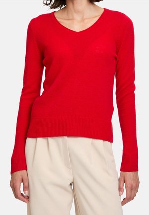 Femme portant un pull rouge ajusté à col en V à manches longues et un pantalon plissé crème taille haute sur fond blanc.