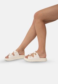 Des sandales blanches à enfiler avec deux brides réglables, des boucles en or et une semelle texturée. Elles présentent un design ouvert au niveau des orteils, une forme moderne et un style minimaliste.