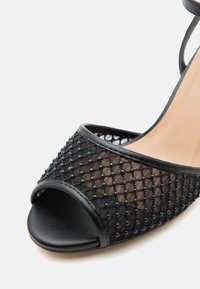 Μαύρο πέδιλο ανοιχτής μύτης με δίχτυ και μικρές στρογγυλές διακοσμήσεις, με λεπτό λουράκι στον αστράγαλο και μπεζ εσωτερική σόλα.