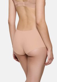 Ženska v nedrčku brez šivov v nude barvi in ujemajočih se hipster hlačkah, prikazana s hrbtne strani na svetli ozadju.