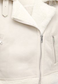 Kurtka w kolorze kremowym z miękkim, teksturowanym kołnierzem z sztucznego shearlingu. Posiada pełny zamek, boczne kieszenie oraz gładką tkaninę.
