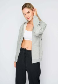 Felpa grigia con zip, dal tessuto morbido, con interno foderato e due tasche frontali, indossata sopra un top corto bianco e pantaloni neri con coulisse.