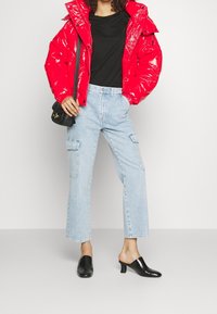 Veste matelassée rouge brillant, t-shirt noir, jeans cargo taille haute bleu clair, et mules à talons noirs complètent la tenue.