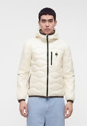 Jeune homme portant une veste matelassée crème avec des détails de fermeture éclair noirs et un jean bleu clair, debout devant un fond blanc uni.