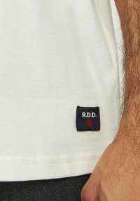 Short-sleeve, cream cotton t-shirt with a navy fabric label featuring "R.D.D." and a red dot accent near the hem.