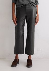Schwarze Leder-Hosen mit weitem Bein, glatter Textur und sichtbaren Nähten, kombiniert mit braunen Wildleder-Schlipschuhen.