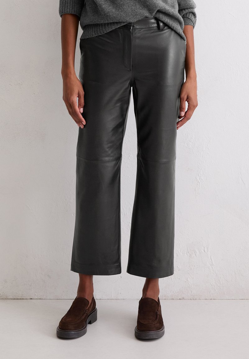 Schwarze Leder-Hosen mit weitem Bein, glatter Textur und sichtbaren Nähten, kombiniert mit braunen Wildleder-Schlipschuhen.