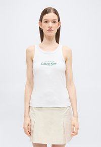 Ung kvinde med glat brunt hår iført en hvid, ærmeløs Calvin Klein-top og en beige nederdel, stående mod en ensfarvet baggrund.