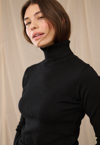 Mulher com cabelo castanho curto, vestindo um suéter de gola alta preto, olhando ligeiramente para cima com um fundo bege neutro.