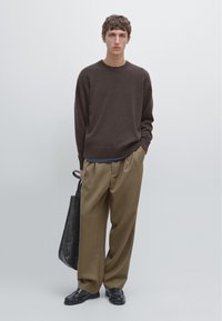 Brauner gestrickter Pullover, lockere beige Hose und schwarze Lederschuhe. Das Modell hält eine schwarze Tragetasche. Einfaches, minimalistisches Design.