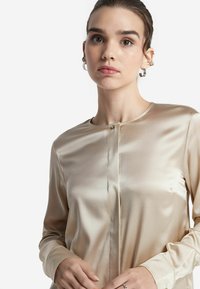 Silkesblus i ljusbeige med långa ärmar, rund halsringning och framknäppning. Slenyt och diskret lyster.