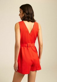 Combinaison en lin rouge avec un dos en V, une conception sans manches et une taille ceinturée. Les jambes courtes et un tissu texturé complètent le look.