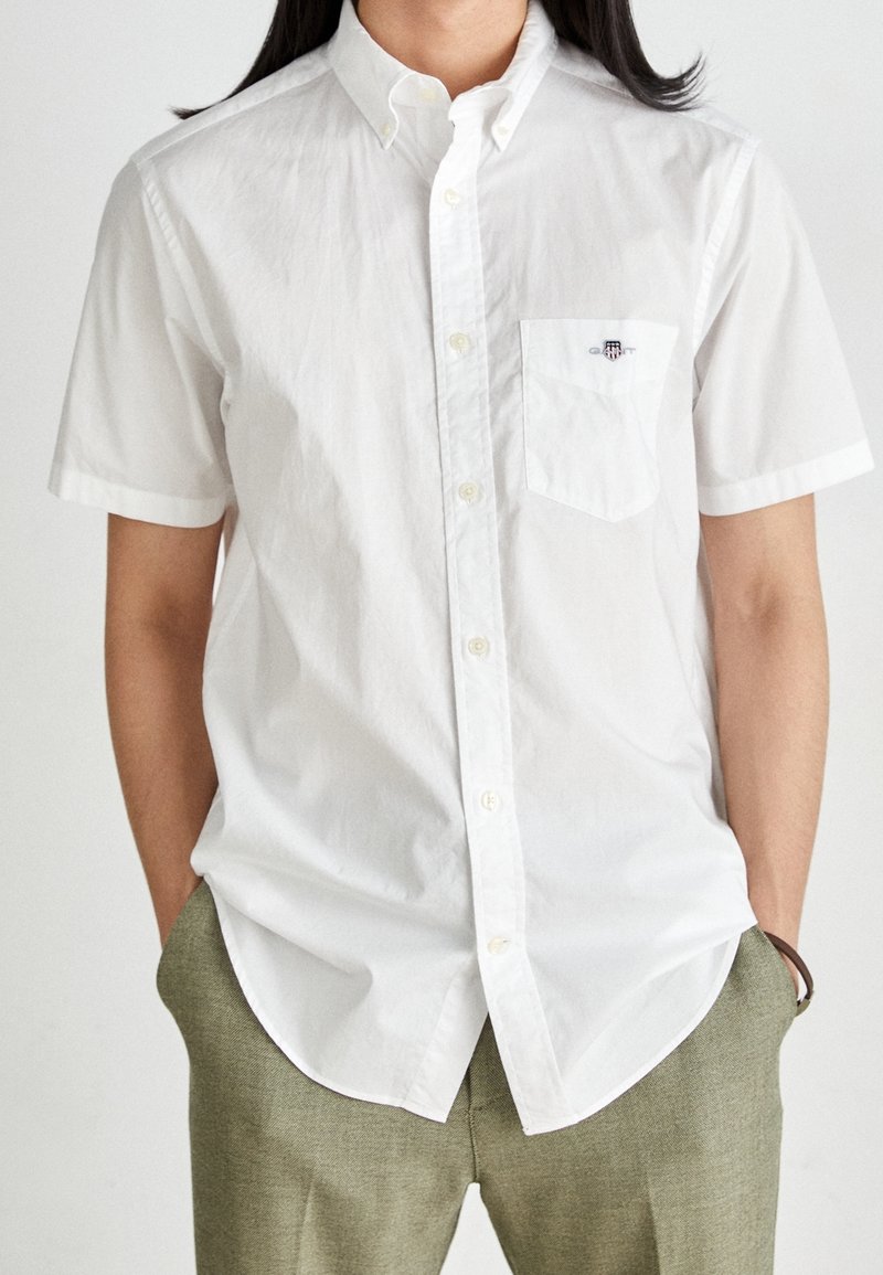 Personne portant une chemise blanche à manches courtes avec un petit logo brodé sur la poche poitrine et un pantalon vert olive.