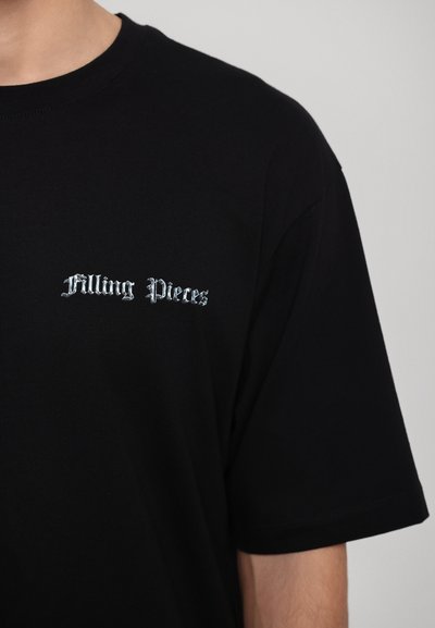 Melnā kokvilnas t-krekls ar īsām piedurknēm, kuram priekšā ir "Filling Pieces" logo sudraba gotiskajā fontā. Gluda tekstūra, klasiska piegriezuma.