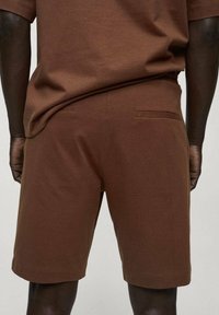 Homme portant un short en maille marron et un haut à manches courtes marron assorti, vu de dos sur un fond uni.