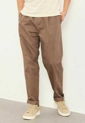 Man wearing brown casual pants with rolled cuffs and beige sneakers standing against a light wall with hands in pockets.