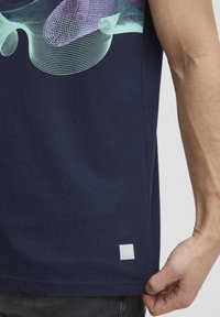 T-shirt in cotone blu navy con un motivo geometrico in azzurro chiaro e viola, dotato di un piccolo etichetta bianca sull'orlo.