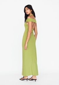 Robe verte décolletée avec un corsage ajusté et un dos ouvert. Texture lisse, longueur au sol, assortie à des sandales à talons noires à lanières.