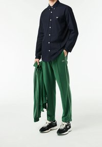 Camicia a bottoni navy, pantaloni tuta verdi con strisce bianche ai lati, mentre tiene un giubbotto verde abbinato e sneaker nere con dettagli bianchi.
