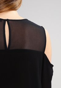 Svart skjorta med genomskinlig övre sektion och detaljer med nyckelhål på baksidan. Har mjukt, texturerat tyg och axelkotsar för en lager-på-lager-look.