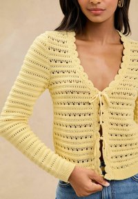 Adīta dzeltena kardigāna ar viļņotām kakla malām, dekoratīvām saitēm un horizontālu svītru tekstūru. Iezīmējas ar atvērta darba dizainu un pieguļošām piedurknēm.