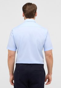 Camicia azzurro chiaro a maniche corte con colletto diritto, texture liscia, spacco posteriore e senza motivi visibili, abbinata a pantaloni blu navy.