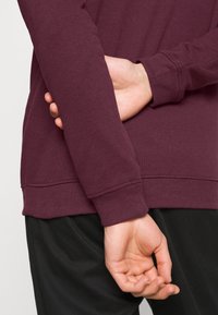 Person iført en mørkerød langærmet skjorte med den ene hånd, der griber om det modsatte håndled bag ryggen, iført sorte bukser.