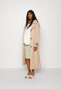 Casaco trench bege com cintura cingida, grandes lapelas e detalhes com botões, usado sobre uma blusa branca e uma saia riscada verde.