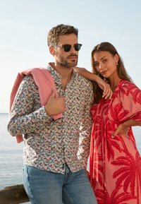 Lichtgepatroneerde, languitgevoerd blouse met bloemenmotief, gedragen met blauwe jeans. Model draagt een zonnebril en houdt een roze trui vast. Heldere rode jurk met palmenbladmotief.