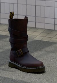 Σκούρο καφέ δερμάτινη μπότα μέχρι τη μέση της γάμπας με περιμετρικούς ιμάντες, ορειχάλκινη αγκράφα, χοντρή σόλα με κίτρινο ράψιμο, πάνω σε πεζοδρόμιο κοντά σε τοιχο με πλακάκια.