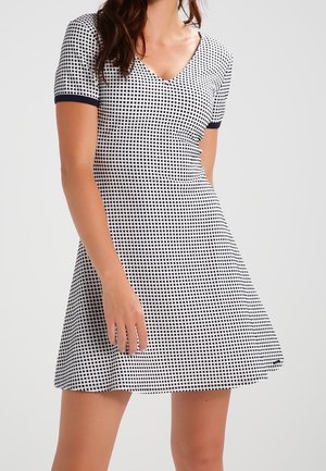 Nainen, joka seisoo yksivärisen taustan edessä ja pukeutunut lyhythihaiseen, mustavalkoruudulliseen mekkoon, jossa on V-aukko ja istuva vyötärö.