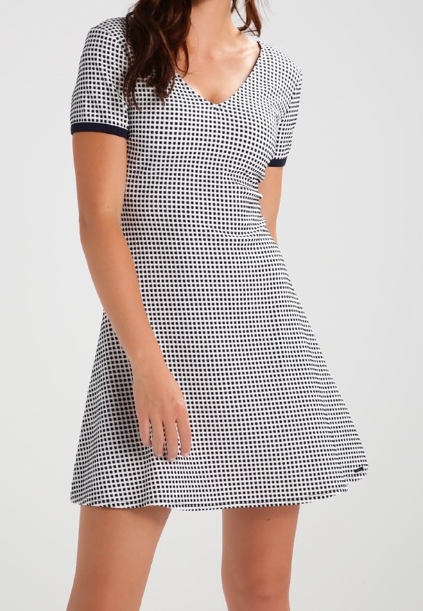 Femme portant une robe à manches courtes à carreaux noir et blanc avec un décolleté en V et une taille cintrée, debout devant un fond uni.
