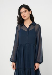 Μπλε ναυτικό φόρεμα κατασκευασμένο από ελαφρύ, διαφανές ύφασμα. Διαθέτει εμπρός με κουμπιά, μακριά μανίκια φουσκωτά και φούστα με πολλαπλά στρώματα.