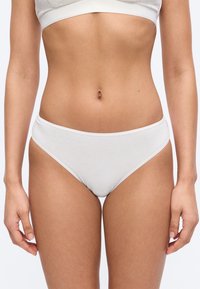 Γυναικείο σώμα με λευκά βαμβακερά εσώρουχα και λευκό στηθόδεσμο, που στέκεται μπροστά σε απλό ανοιχτόχρωμο φόντο.