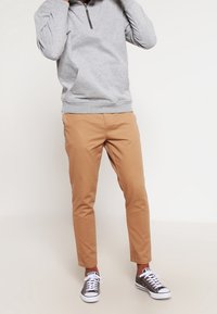 Persona che indossa un maglione grigio chiaro con zip a 1/4, pantaloni beige e sneakers grigie basse, in piedi su uno sfondo bianco semplice.