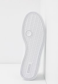 Semelle en caoutchouc blanche avec motif en chevrons, rainure circulaire près de la tête du pied, et logo « LACOSTE » embossé près du talon.
