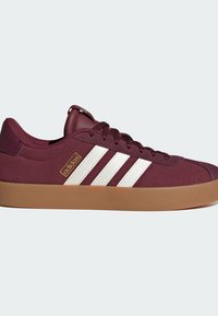 Červené semišové tenisky s bílým designem s třemi pruhy, béžovou gumovou podrážkou a zlatou visačkou s logem. Mají zaoblenou špičku a polstrovaný límec.