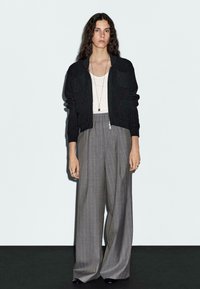 Black textured cardigan with two front pockets, paired with high-waisted gray striped pants. Accessories include a long necklace and rings.