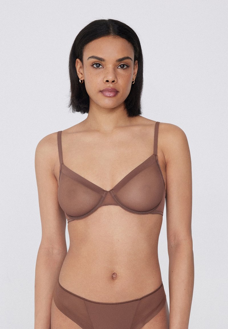 Frau mit kurzen dunklen Haaren, die einen braunen transparenten BH und passende Unterwäsche trägt, vor einem schlichten weißen Hintergrund stehend.