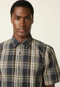 Košeľa s krátkym rukávom v károvanom vzore, obsahujúca tmavomodré, hnedé a krémové farby, golier na zapínanie na gombíky a štruktúrovanú tkaninu.