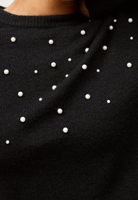 Schwarzer gestrickter Pullover mit verstreuten weißen Perlenverzierungen, der einen strukturierten Stoff und einen Rundhalsausschnitt präsentiert.