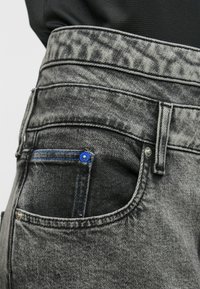 Γκρίζα τζιν με ψηλή μέση, υφή, μπλε ραφές στην πίσω τσέπη και μεταλλικά καρφιά για ενίσχυση.
