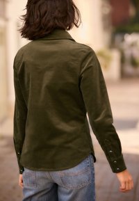 Camicia a coste verde oliva con design sulla spalla posteriore, maniche lunghe e bottoni a pressione ai polsini. Tessuto morbido, vestibilità casual.